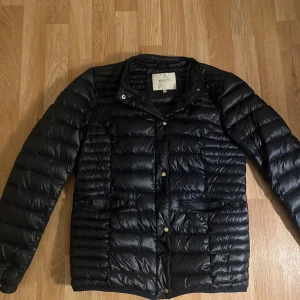 Svart dunjacka från Selected Femme - Snygg svart dunjacka från Selected Femme i storlek 36. Jackan har quiltad design, guldiga knappar och dragkedja framtill. Lätt och smidig modell med lång ärm, perfekt för kyliga dagar. Klassisk look som funkar till många outfits.