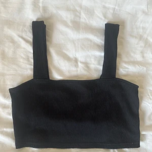Svart ribbad croppad linne - En svart croppad linnetopp med breda axelband och ribbad struktur. Toppen är ärmlös och har en enkel, stilren design som passar perfekt till jeans eller kjol. Materialet är stretchigt och bekvämt, vilket gör den lätt att bära hela dagen