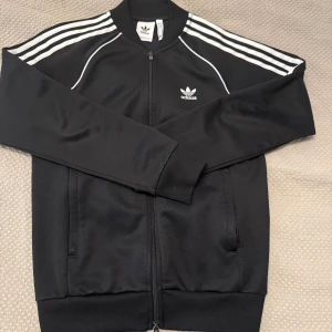 Svart Adidas tröja S  - Klassisk svart Adidas track jacket med vita ränder. Tröjan är i väldigt bra skick och inga fel på den alls.