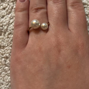 Guldfärgad ring med två pärlor - Snygg guldfärgad ring med två vita pärlor i olika storlekar. Ringen har en öppen design där pärlorna sitter i varsin ände och ger en modern och stilren look. Perfekt för dig som gillar klassiska detaljer med en twist. OBS!!! KOMMER GÖRAS RENT INNAN SICKAD
