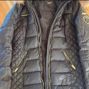 Svart dunjacka Rockandblue med fuskpäls - Snygg svart dunjacka från Rockandblue med quiltade detaljer, guldiga dragkedjor och badge på ärmen. Jackan har en stor huva med fluffig fuskpäls och flera fickor med dragkedja. Perfekt för kalla dagar när du vill vara både varm och stilren. Äkta päls. Bara använd en vinter  