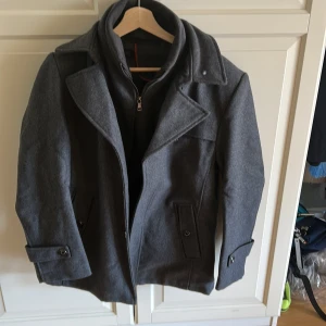 Grå skepparkavaj i ullmix -  Grå skepparkavaj med klassisk krage, knappar framtill och snygga detaljer vid ärmslut och fickor.  Säljes pga av felköp. Storlek L på lappen men är mer en M.