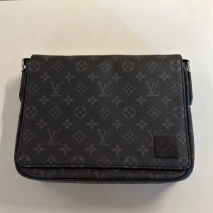 Louis Vuitton svart axelväska monogram - Snygg svart axelväska från Louis Vuitton med klassiskt monogrammönster i grått. Väskan har justerbar axelrem, dragkedjefack på baksidan och detaljer i skinn. Perfekt för dig som vill ha en stilren och lyxig accessoar med ikonisk design.