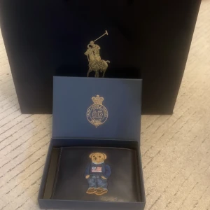 Svart plånbok Polo Ralph Lauren björn - Svart plånbok i skinn från Polo Ralph Lauren med broderad ikonisk björn på framsidan. Plånboken har flera kortfack och präglad logga på baksidan. Perfekt accessoar för dig som gillar klassisk stil med en twist.