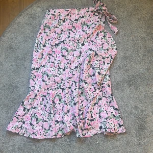 Blommig omlottkjol från Gina Tricot - Supersöt omlottkjol från Gina Tricot i blommigt mönster med rosa, gröna och svarta toner. Kjolen har knyt i midjan och volang nertill för en extra flowy känsla. Perfekt för dig som vill ha en feminin och trendig look.