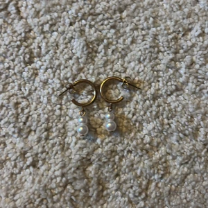 Guldiga örhängen med pärlor - Snygga örhängen i guldfärgad metall med två vita pärlor som hänger i varje ring. Perfekt accessoar för att lyfta din stil med en elegant touch. Runda hoops med klassisk design och pärldetaljer som ger en trendig vibe. OBS!!! KOMMER GÖRAS RENT INNAN SICKAD