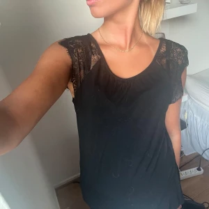 Svart topp med spetsdetaljer - Säljer en svart ärmlös topp med snygga spetsdetaljer över axlarna. Toppen har rund halsringning och ett flowy fall. Perfekt för dig som gillar en enkel men ändå lite extra look. Tröjan är från Hunkemöller 