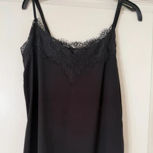 Svart linnetopp med spetsdetaljer - Snygg svart linnetopp med tunna axelband och vacker spets längs urringningen. Perfekt för att skapa en stilren och trendig look. Toppens material känns mjukt mot huden och spetsen ger en extra touch av elegans.