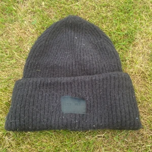 Svart ribbstickad mössa one size - Svart ribbstickad mössa med uppvikt kant och diskret patch framtill. Tillverkad i en mjuk blandning av akryl, polyamid och elastan som ger både värme och stretch. Enkel och stilren design som passar till det mesta.