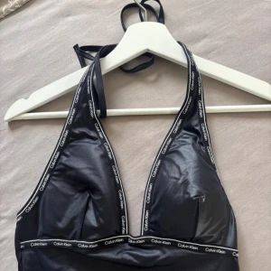 Svart baddräkt från Calvin Klein - Snygg svart plunge baddräkt från Calvin Klein med logotypband längs kanterna. Djup urringning och öppen rygg ger en modern look. Tillverkad i glansigt syntetmaterial som sitter tight och bekvämt. Perfekt för stranden eller poolen.