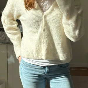 Vit stickad kofta från American Vintage - Mysig vit stickad kofta från American Vintage med rund halsringning och knäppning framtill. Koftan har långa ärmar och en mjuk, fluffig känsla. Perfekt att slänga över en t-shirt eller linne för en avslappnad look.