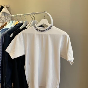 Vit t-shirt med logga från Acne Studios - Vit t-shirt från Acne Studios med svart logotyptext runt den ribbade halsringningen. Klassisk passform och korta ärmar. Storlek XS, unisex. Har en fläck som inte går bort, se bild 3 dör referens. Däremot så syns inte fläcken särskilt mycket, annars fint skick. Pris kan diskuteras! 