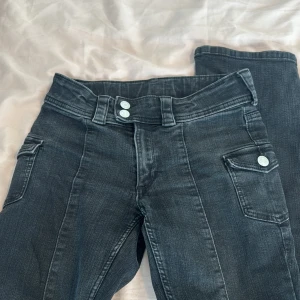 Svarta bootcut jeans från H&M - Säljer ett par svarta bootcut jeans från H&M med låg midja och justerbar midja. Jeansen har fickor med lock och silverfärgade knappar framtill. Perfekta för dig som gillar en klassisk men cool stil.
