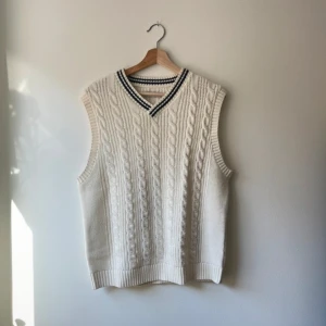 Vit stickad väst med kabelmönster från Jack & Jones - Vit stickad väst från Jack & Jones med klassiskt kabelstickat mönster och v-ringad hals med svarta ränder. Västen har en normal passform och är tillverkad i en blandning av akryl, ull och bomull.