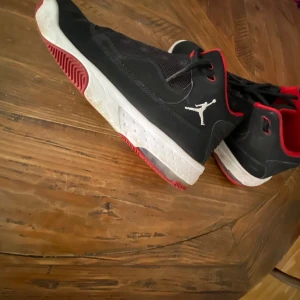 Svarta sneakers från Jordan med röd och vit sula - Säljer ett par svarta Jordan sneakers med vit och röd sula. Skorna har den klassiska Jumpman-loggan på sidan och detaljer i rött runt öppningen. Perfekta för dig som gillar sportig stil.