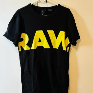 Svart t-shirt med RAW-tryck från G-Star RAW - Cool svart t-shirt från G-Star RAW med stort gult RAW-tryck på framsidan. Klassisk passform och rund halsringning. Perfekt för dig som gillar streetwear och vill sticka ut med en enkel men snygg look.