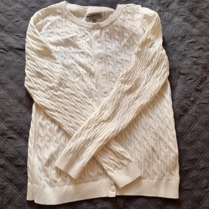 Vit kabelstickad kofta från Basic You - Säljer en vit kabelstickad kofta från Basic You i ekologisk bomull. Koftan har rund halsringning, knappar framtill och långa ärmar. Perfekt för lager-på-lager och enkel att matcha med olika outfits. Aldrig använt! Pris kan diskuteras!