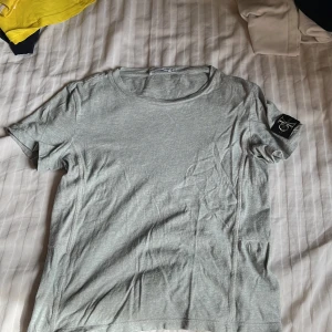 Ljusgrå t-shirt från Calvin Klein Jeans - Säljer en ljusgrå t-shirt från Calvin Klein Jeans med rund halsringning och korta ärmar. T-shirten har en stor svart CK-logga på ärmen och är tillverkad i mjuk bomull. Perfekt för en avslappnad och stilren look.