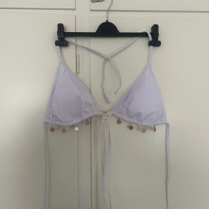 Vit bikiniöverdel med gulddetaljer - Säljer en vit trekantsbikini med knyt i nacken och ryggen. Bikinin har små guldfärgade myntdetaljer längs nederkanten som ger en unik touch. Den har ett snöre man kan knyta i revbenen för en snyggare bikini.