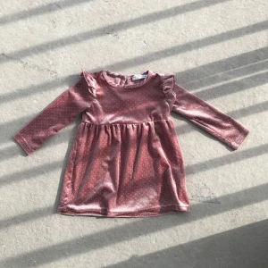 Rosa sammetsklänning med volang och prickar - Superfin rosa sammetsklänning med små prickar och volangdetaljer vid axlarna. Klänningen har lång ärm och rund halsringning, perfekt för en söt och trendig look.