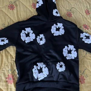 Denim tears helt oanvänd  - Säljer en svart hoodie med stora vita blommor tryckta över hela tröjan, inklusive huvan och fickan. Hoodien har en klassisk passform och en stor magficka framtill. Perfekt för dig som vill sticka ut med en unik och snygg stil.