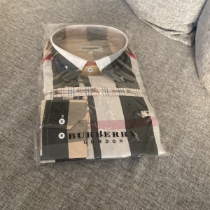 Rutig skjorta från Burberry London - Säljer en klassisk rutig skjorta från Burberry London i storlek L. Skjortan har långa ärmar, knappar framtill och ett ikoniskt mönster i beige, svart, vit och röd. Perfekt för dig som gillar stilrena och tidlösa plagg.
