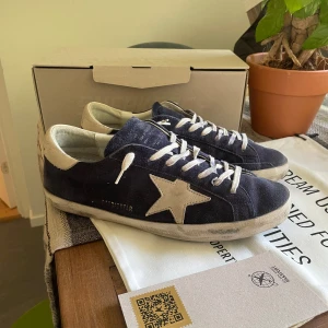 Mörkblå sneakers från Golden Goose med vit stjärndetalj - Säljer ett par mörkblå sneakers från Golden Goose med vit stjärna på sidan och vita snören. Skorna har en klassisk låg modell och är tillverkade i mocka med detaljer i vitt skinn. Perfekta för dig som gillar en stilren men unik i använt skick 