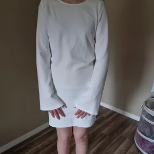 Vit långärmad kortklänning - Säljer en stilren vit klänning med lång ärm och rund halsringning. Klänningen är kort och har en snygg drapering i ryggen med knytband. Storlek m och ny