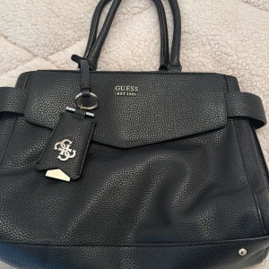 Svart handväska från Guess - Säljer en stilren svart handväska från Guess med silverdetaljer och logotyp framtill. Väskan har flera fack, både med dragkedja och magnetlås, samt ljus insida med Guess-tryck. Perfekt för att bära med sig det viktigaste.