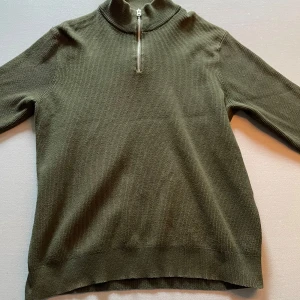 Olivgrön stickad tröja med halv dragkedja från Only & Sons - Säljer en olivgrön stickad tröja från Only & Sons med hög krage och halv dragkedja. Tröjan har lång ärm och är ribbstickad vid krage, ärmslut och nederkant. Perfekt för lager-på-lager under kyliga dagar.