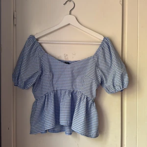 Blåvit rutig blus med puffärmar - Supersöt blus i blåvitt rutigt tyg med puffiga korta ärmar och volang nertill. Sitter snyggt offshoulder och har en romantisk vibe. Perfekt till jeans eller kjol för en somrig look.