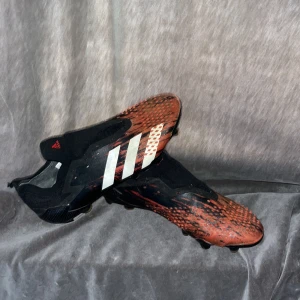 Adidas fotbollsskor elite - Säljer ett par Adidas fotbollsskor i svart och rött med vita ränder på sidan. Skorna har en låg profil och är tillverkade i syntetmaterial, perfekta för spel på gräsplan. Snygg och modern look med mönstrad ovandel.