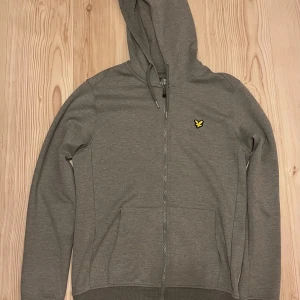 Grå hoodie från Lyle & Scott - Säljer en stilren grå hoodie från Lyle & Scott med dragkedja och huva. Tröjan har ett litet broderat logomärke på bröstet och ribbade muddar vid ärmslut och nederkant. Perfekt för en avslappnad look.