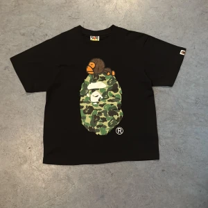 🐵BAPE T SHIRT CAMO🐵 - Svart t-shirt från BAPE med stort camo-tryck av apans ansikte på bröstet och en liten apa ovanpå. Klassisk rund hals och korta ärmar. Ikonisk BAPE-logga på ärmen och etikett i nacken.