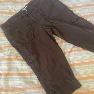 Bruna cargopants från XLNT Woman - Bruna cargopants från XLNT Woman med flera fickor och knappdetaljer på benen. Byxorna har en avslappnad passform och är tillverkade i bomull. Perfekta för en chill och bekväm stil. Saknas en knapp på fickan därbak se bild 5