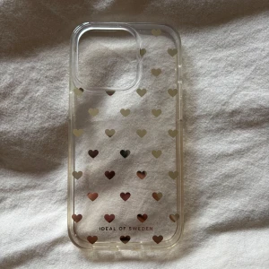 Transparent mobilskal med hjärtan från iDeal of Sweden - Säljer ett transparent mobilskal från iDeal of Sweden med små guldiga hjärtan över hela skalet. iPhone 14 Pro