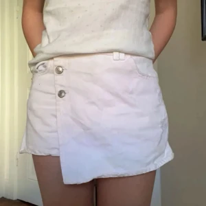 Vit jeanskjol med knappar - Säljer en vit jeans kortkjol med inbyggda shorts och asymmetrisk nederkant. Kjolen passar perfekt till sommaren. Enkel och fräsch design.