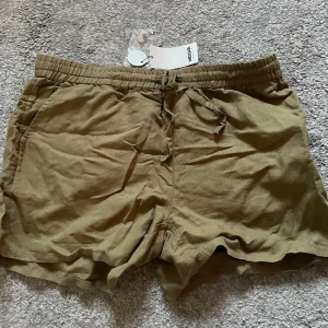 Olivgröna shorts från Indiska - Säljer ett par olivgröna shorts från Indiska med resår i midjan och snörning framtill. Shortsen har två fickor fram och två bakfickor med knapp. Linne.