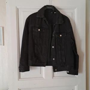 Svart jeansjacka från KappAhl - Klassisk svart jeansjacka med silverfärgade knappar och två bröstfickor. Rak passform och krage, perfekt att styla till olika outfits. Jackan har långa ärmar och tydliga sömdetaljer för en tidlös look.