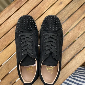Svarta Louboutin med spikar - Säljer ett par svarta sneakers från Christian Louboutin med svarta nitar på tån och ovandelen. Skorna har klassisk röd sula, snörning och är tillverkade i mocka. Perfekta för dig som vill sticka ut med en edgy och lyxig stil.