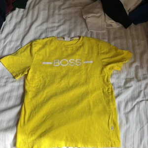 Gul t-shirt från BOSS - Säljer en gul t-shirt från BOSS med vit logga tryckt på bröstet. Klassisk rund halsringning och korta ärmar. Perfekt för dig som vill sticka ut med en färgglad och stilren look.