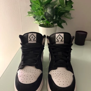 Svarta och vita Nike Air Jordans - Säljer ett par klassiska Nike Air Jordans i svart och vitt. Skorna har svart snörning, svart swoosh och detaljer i mocka samt perforerad tå. Mellanhöga med rund tå och platt sula. Storlek 38