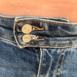 Blå bootcut jeans med råa kanter och dubbelknäppning - Säljer ett par blå bootcut jeans från lager157 med låg midja och smal passform. Jeansen har förlängt midjeband med dubbla knappar och dragkedja i gylfen. Råa, slitna kanter vid bensluten. Gjorda i comfort stretch med fickor fram och bak. Jeansen har skada längst ner på byxorna med också lite vid knappen på byxorna. Annars är de i relativt nyskick då jag köpte de för endast 2,5 månader sedan. Katt finns i hemmet.  Nypris 400kr.