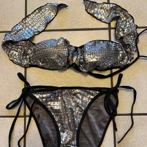Glittrig silver bikini med knyt - Säljer en snygg bikini i glittrigt silver med reptilmönster. Överdelen har knyt i nacken och baktill, och bikinitrosan har knyt i sidorna. Perfekt för dig som vill sticka ut på stranden!