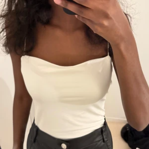 Vit bodysuit med tunna axelband - Säljer en stilren vit bodysuit med tunna axelband och lätt draperad urringning. Perfekt att matcha med både jeans och kjol för en trendig look. Den har en slät och figurnära passform.
