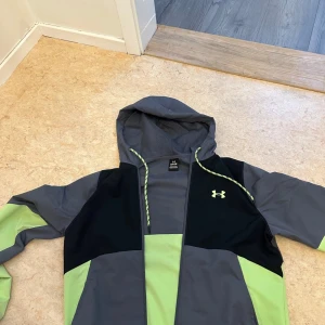 Grå och svart jacka från Under Armour - Säljer en grå och svart jacka från Under Armour med gröna detaljer. Jackan har en huva och dragkedja framtill. Perfekt för träning eller vardagsbruk. Den har långa ärmar och en sportig look.