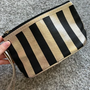 Randig necessär i guld och svart - Snygg necessär med breda svarta och guldiga ränder. Den har dragkedja upptill och ett litet handtag i guld. Insidan är ljus och enkel att rengöra.