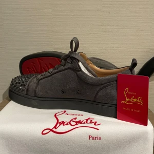 Svarta sneakers med nitar från Christian Louboutin - Säljer ett par svarta sneakers från Christian Louboutin med mattsvarta nitar på tån och runt sulan. Skorna har snörning och är tillverkade i mocka med en platt sula. Riktigt unika och sticker ut i mängden.