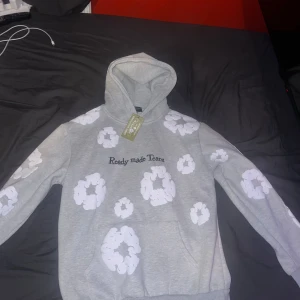 Grå denim tear hoodie - Säljer en grå hoodie från denim tear med vita blommor tryckta över hela tröjan och texten 'Ready-made Tears' på bröstet. Tröjan har huva och är i mjukt material, perfekt för en avslappnad stil. Storlek S men kan även passa M