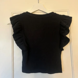 Svart ribbad topp med volangärmar från Zara - Supersnygg svart topp från Zara med ribbat material och volangdetaljer på ärmarna. Modellen är tight och passar perfekt till många olika outfits. Enkel rund halsringning och korta ärmar med extra volym.